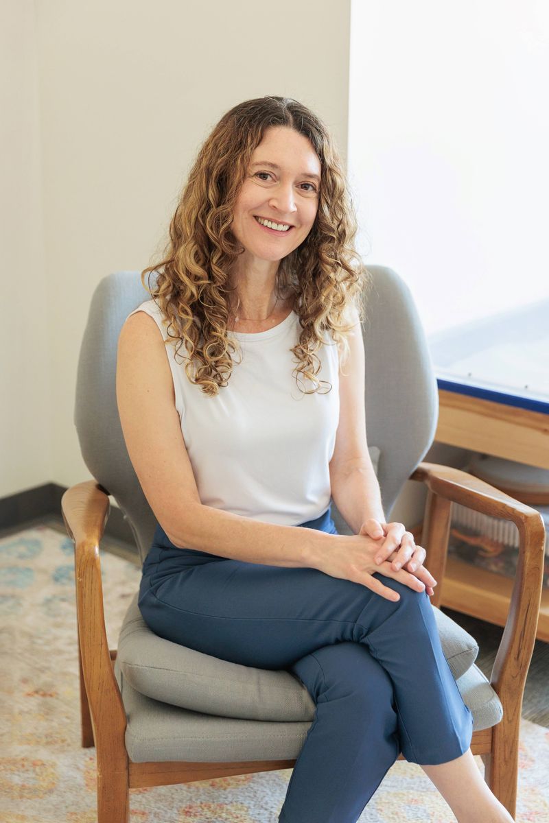 Miché Jo-Keeling, MSW RSW. Her long wavy brown hair touches below her shoulders and she wears a zippered coat. The background is a soft green.